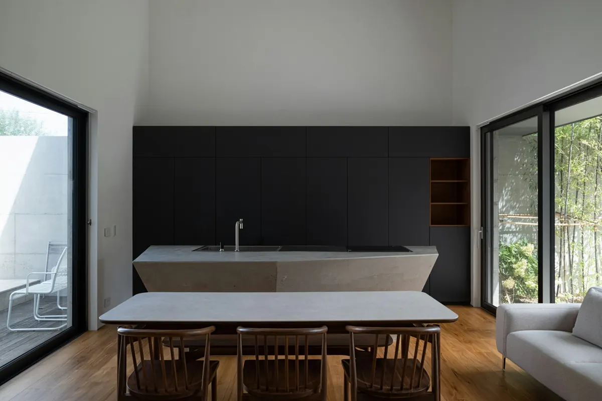 A long wall of matte black full-height cabinetry behind a pale faceted stone kitchen island with a sink and a flush cooktop, a wooden dining table and chairs in the foreground, glass doors to a garden on both sides.