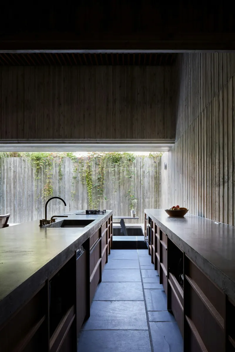 A narrow kitchen with two long concrete-topped counters over dark timber drawers, a slate floor between them, timber-clad walls, and a wide opening at the end framing a fence covered in climbing plants.