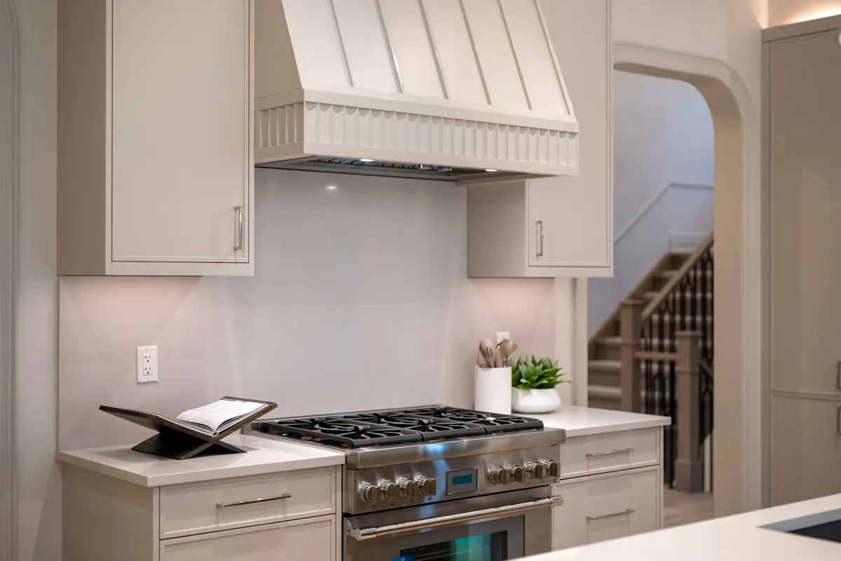 A custom painted range hood with a fluted lower band above a six-burner stainless range, flanked by greige cabinets, with a staircase visible through an arched opening.