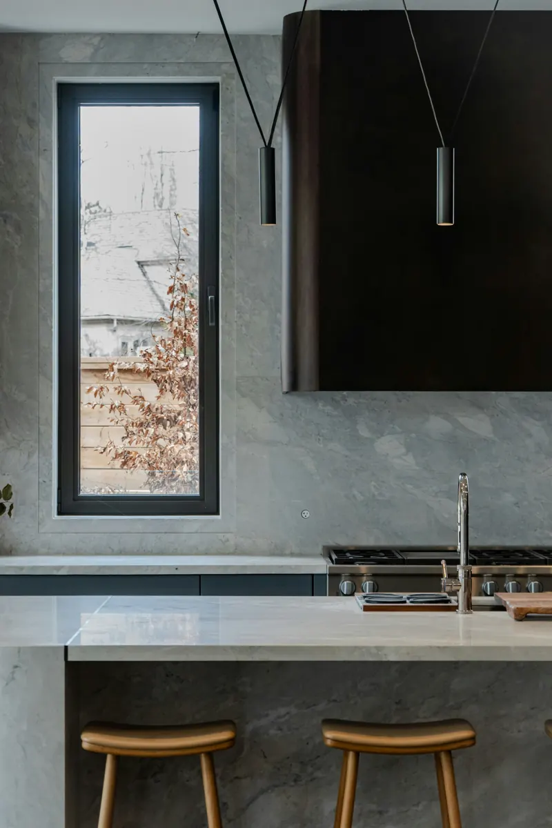 A tall kitchen window beside a wall of grey veined stone, with a dark walnut range hood above a stainless pro-style range and a stone island with two wooden stools.