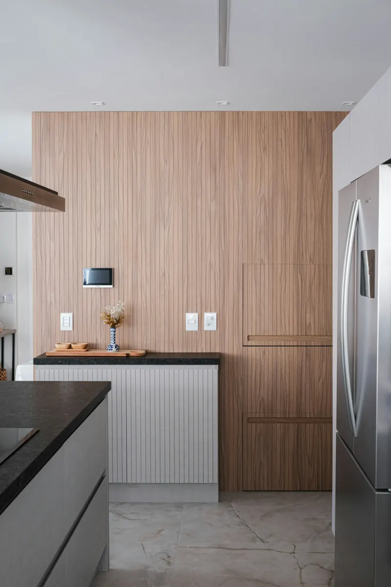 A kitchen wall clad in vertical walnut paneling with flush panel doors and recessed pulls, a white fluted counter cabinet with a dark stone top, and a stainless French-door refrigerator standing at the right edge.
