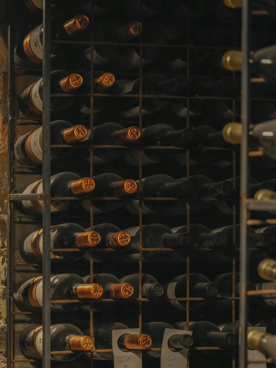 Rows of wine bottles with copper-colored capsules resting on their sides in an iron rack against a stone wall, a few handwritten tags hanging below.